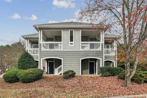 A grey two-story house with a balcony and a tree with red leaves in front.