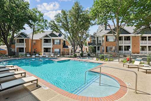 A swimming pool surrounded by lounge chairs and trees in front of apartment buildings.