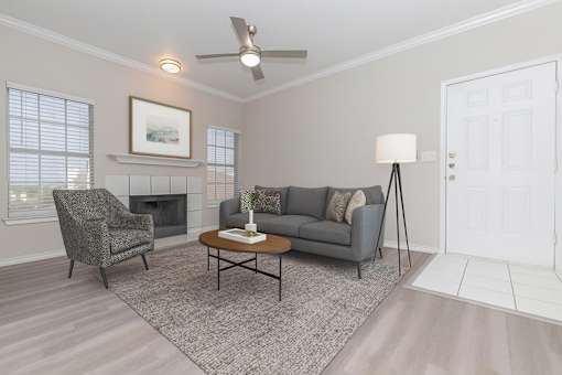 A living room with a grey couch, a grey chair, a grey floor lamp, a grey rug, a grey coffee table, a grey fireplace, a white door, a white wall, and a white ceiling fan.