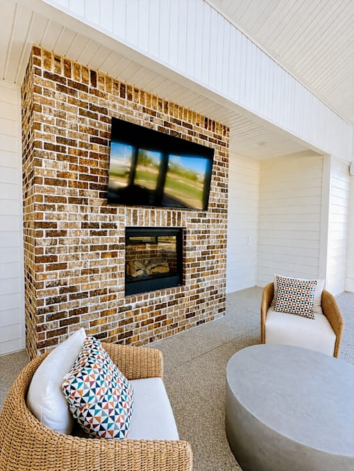 A wicker chair with a cushion sits in front of a brick fireplace with a television above it.