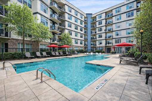 A pool surrounded by chairs and umbrellas in front of apartment buildings.
