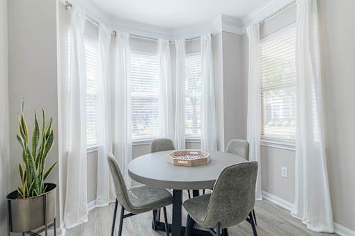 A round table with four chairs and a potted plant in a room with white curtains.