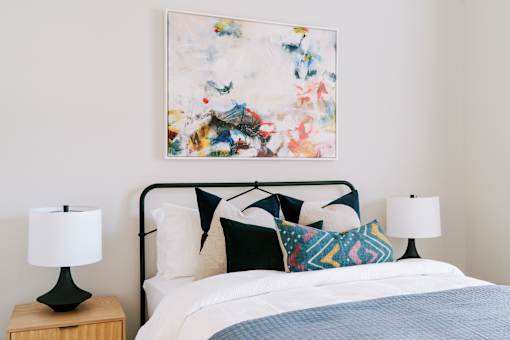 A bed with a white comforter and a blue blanket with a white pillow and a painting on the wall.