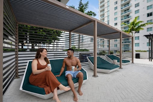 A man and a woman are sitting on a bench under a shaded area.
