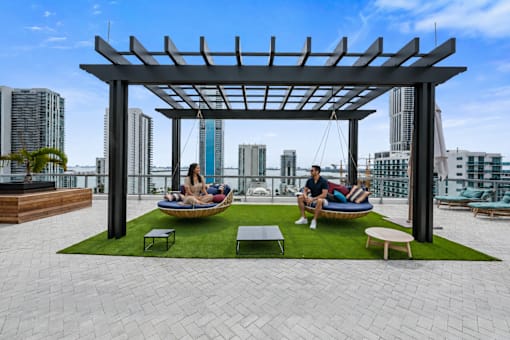 A man and woman are sitting on a couch under a pergola on a rooftop patio.