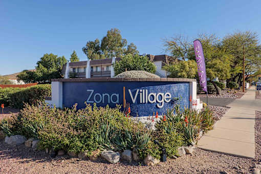 A sign for Zona Village is surrounded by plants and rocks.