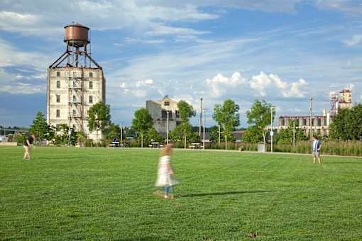 A person in a white dress is running across a grassy field with a large grain silo in the background.