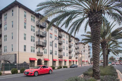 A red car is parked on the side of a street in front of a building with palm trees.