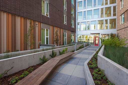 A long, narrow walkway with a bench and a planter on either side leads to a building with a red door.