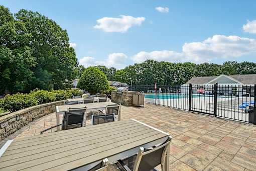 A wooden table and chairs are on a patio with a pool and trees in the background.