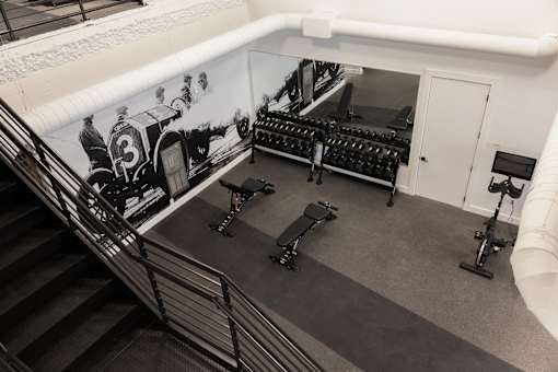 Elevated view of the Cole Motor Apartments fitness center in Indianapolis showing dumbbell racks, weight benches, a stationary bike, and vintage race car mural.