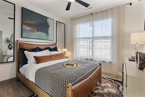 Model apartment bedroom at Montage West Lafayette featuring a modern bed frame, layered bedding, ceiling fan, and large window with natural light.