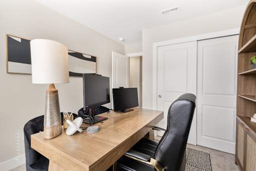 Functional home office space at Mural Lewis Center featuring wood desk, computer setup, ergonomic chair, table lamp, and neutral walls for a productive work environment