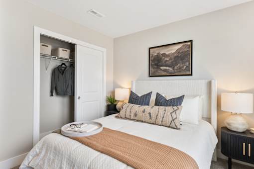 Comfortable bedroom in single-family rental home at Mural Lewis Center featuring plush bedding, neutral decor, bedside lighting, wall art, and walk-in closet with sliding door