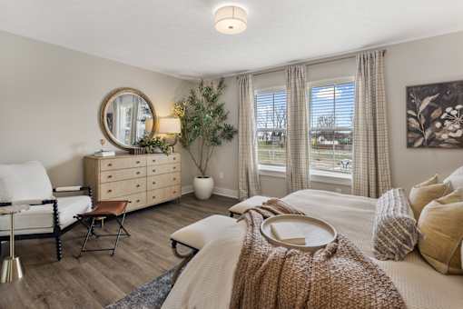 Sunlit primary bedroom with large double windows, plaid drapes, natural wood dresser with round gold mirror, olive tree accent, white upholstered accent chair with leather footstool, botanical wall art, and cozy layered bedding in warm neutral tones