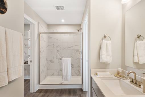 Spacious apartment bathroom at Mural Lewis Center featuring glass-enclosed walk-in shower, double sink vanity, large mirror, modern lighting, and wood-style flooring