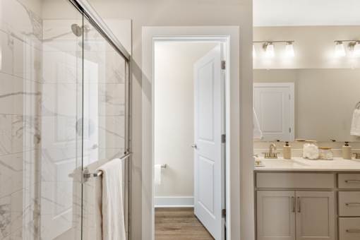Modern bathroom in single-family rental home at Mural Lewis Center featuring glass walk-in shower, dual sink vanity, bright lighting, and wood-style flooring