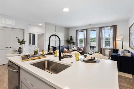 Spacious apartment layout showing a modern kitchen with a large island and a dining area in the background, seamlessly flowing into a bright, sunlit living room.