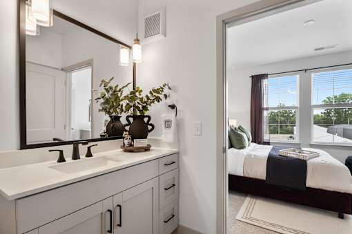 A view from a modern bathroom vanity featuring a light wood cabinet and quartz countertop, looking through an open doorway into a bright, carpeted bedroom with large windows.