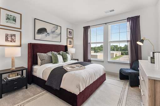A beautifully staged apartment bedroom featuring a premium dark red tufted bed frame, stylish modern nightstands, crisp neutral carpeting, and large windows with elegant drapery that let in bright, natural light.