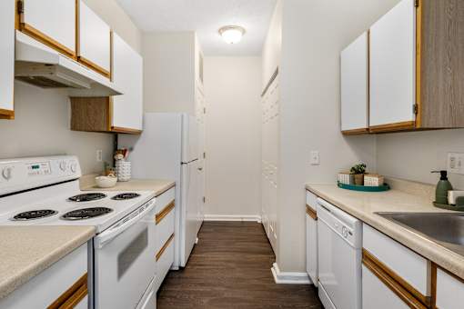 Functional galley-style apartment kitchen featuring white appliances, ample cabinet storage, generous counter space, and wood-style flooring at Steeplechase at Shiloh Crossing Apartments in Avon, Indiana.