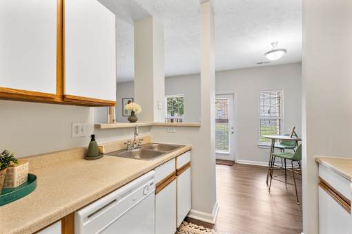 Bright open-concept apartment kitchen featuring white cabinetry, ample countertop space, a double-basin sink, breakfast bar, dishwasher, and luxury vinyl plank flooring flowing into the dining and living areas at Steeplechase at Shiloh Crossing Apartments in Avon, Indiana