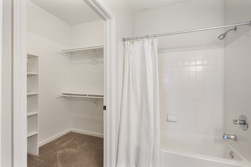 Bright apartment bathroom featuring a full-size bathtub and shower, plus an adjoining walk-in closet with built-in shelving and hanging space at Steeplechase at Shiloh Crossing Apartments in Avon, Indiana.