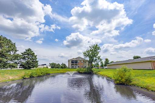 Scenic community pond with a decorative fountain, mature trees, and beautifully landscaped green space at Steeplechase at Shiloh Crossing Apartments in Avon, Indiana