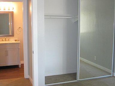 An empty bedroom with tan carpets and a sliding mirror doors to the closet. The bathroom is barely visible to the right.