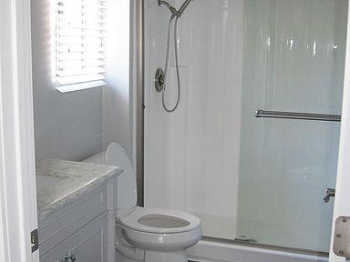 The bathroom with a tub & shower combo that has glass doors. The toilet is in front with a window of light behind and a sink on the left.