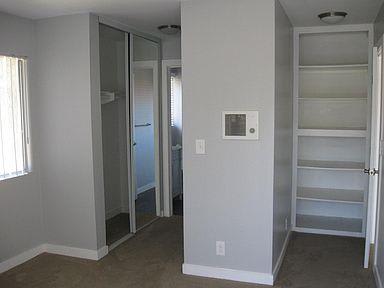A large empty bedroom looks down the hallway lined with closets to the entryway into the bathroom. Shelving is on the right side where the door opens to the bedroom.