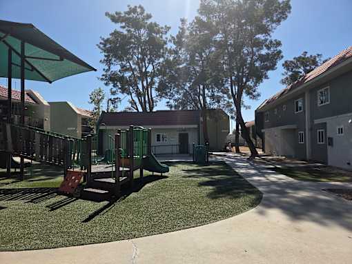A playground with a slide and a green canopy is surrounded by trees and buildings.