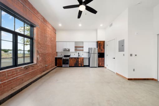 an empty living room with a brick wall and a kitchen with a ceiling fan