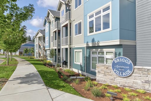 Apartments for Rent in Olympia - Briggs Village - Exterior Apartments Building with Grey and Blue Siding, Lush Greenery, and Sidewalk