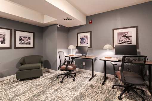 A room with grey walls and a carpeted floor with a green armchair, two desks with chairs, and framed pictures on the wall.