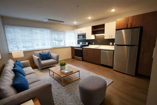 A modern living room with a grey couch, a coffee table, and a kitchen area in the background.