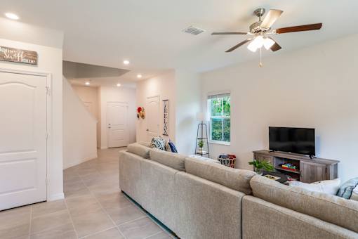 A cozy living room with a large sectional sofa, a ceiling fan, a TV on a wooden stand, and a staircase leading to the upper floor.