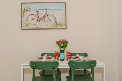 A dining table with two green chairs and a painting of a bicycle on the wall.