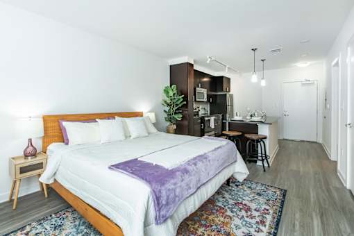 A bedroom with a bed, nightstand, and a kitchen area in the background.