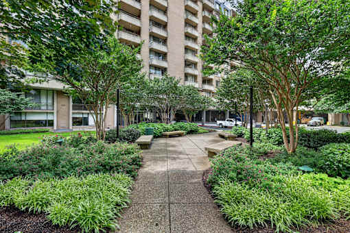 A concrete walkway leads through a landscaped area with a large building in the background.
