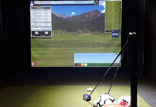 A golf simulator setup with a screen showing a driving range and golf clubs on the floor.