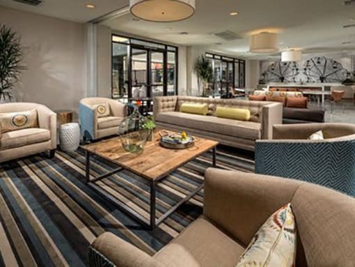 Modern Apartments for Rent in Long Beach, CA - Urban Village - Social Lounge with Carpet Flooring, Sofa, Barrel Armchairs, Wooden Coffee Table, and Large Windows.