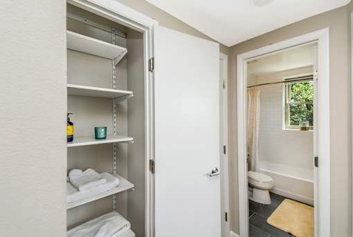 A white bathroom with a white door and a white shelving unit with towels and a bottle.