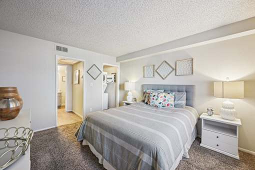 Two-Bedroom Apartments in Mesa, AZ - Mesa Station Apartments - Bedroom With Gray Bedding, Upholstered Headboard, Nightstands, Table Lamps, Wall Decor, Carpeted Flooring, And Laundry Room Access.