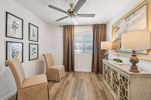 A room with a ceiling fan, two chairs, and a table with a lamp on it.