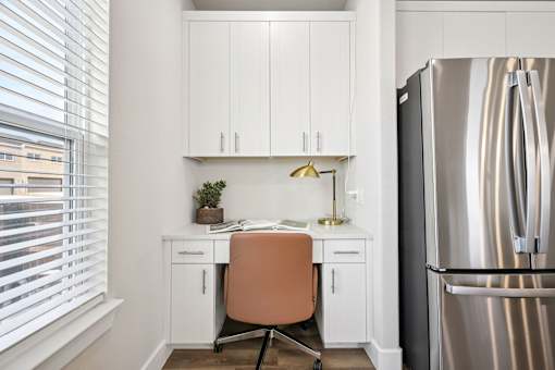 A kitchen with a desk and a chair in front of a refrigerator.
