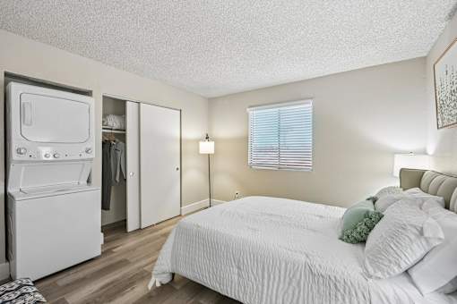 A white bedroom with a bed, a refrigerator, and a window with blinds.