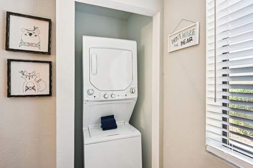 A white washing machine is in a small laundry room with a sign that says "Don't Make the Bear Sad" on the wall.