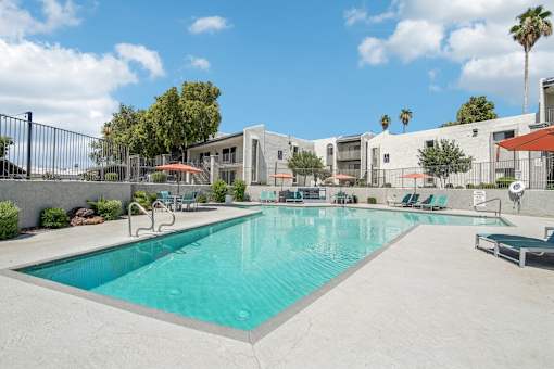 1-Bedroom Apartments in North Mountain Village, Phoenix, AZ - Ardella on 28th - Resort-Style Swimming Pool With Sundeck, Lounge Chairs, Umbrellas, Trees, and Apartment Buildings Under Blue Sky.