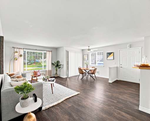 A living room with a grey sofa, a coffee table, a dining table with chairs, and a kitchen area with a countertop.